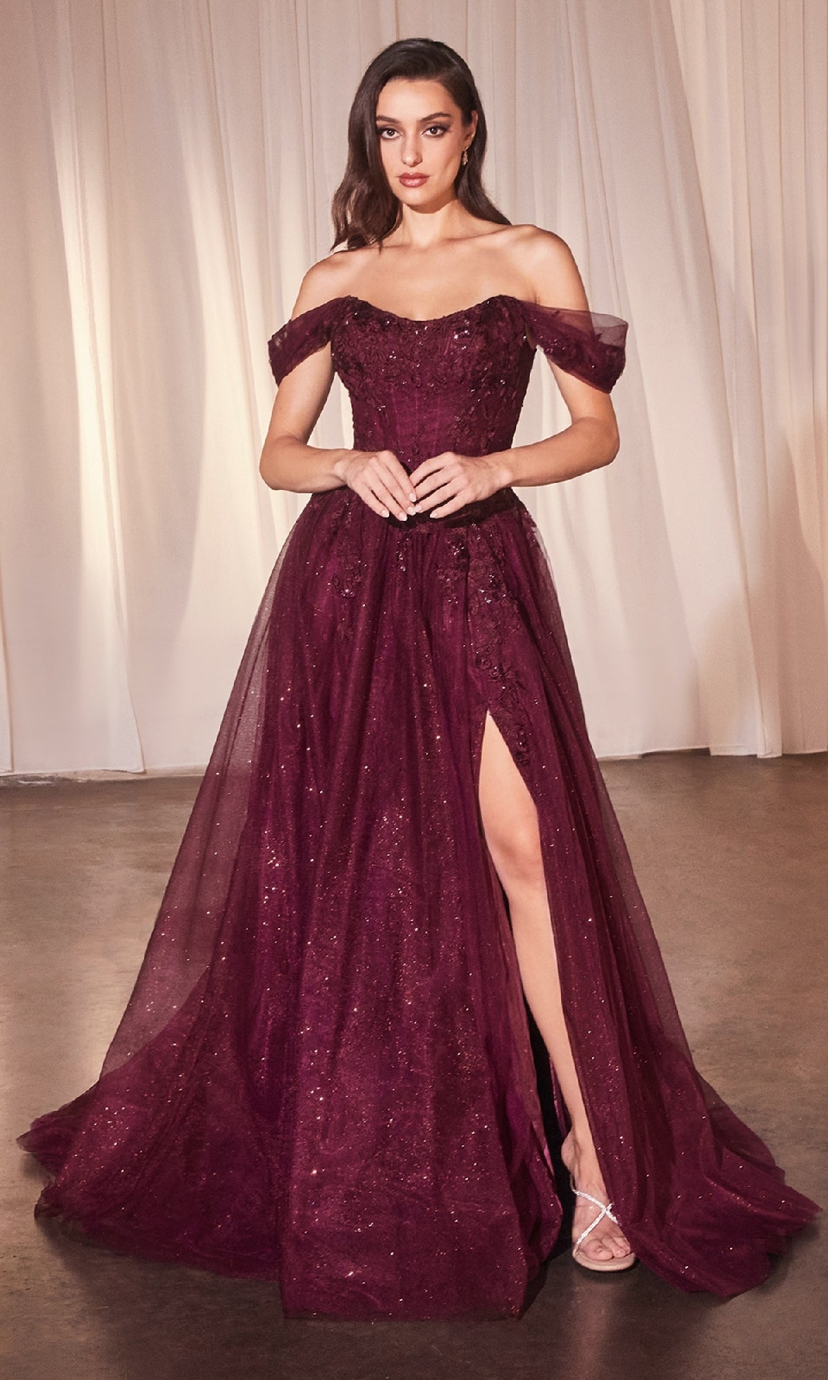 A woman wears the Ladivine CD799 off-shoulder long prom dress in burgundy, featuring a thigh-high slit and glitter detail, standing before white curtains.